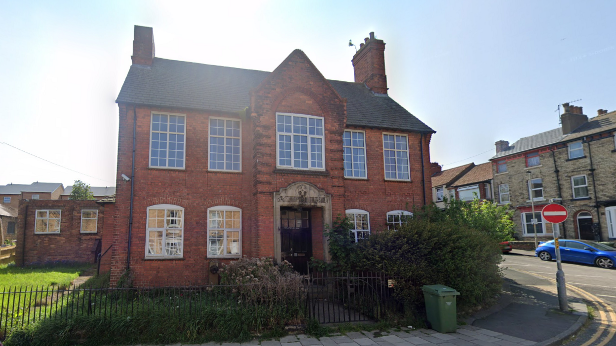 A red-brick building with nine large windows on the front with white edges. The building is on a road with a black metal fence surrounding it. The building has a large blue door.