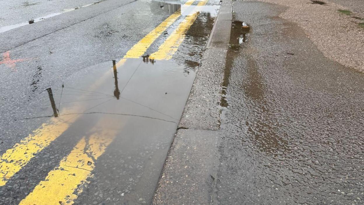 A large puddle in the road over some double yellow lanes, with a pavement alongside.