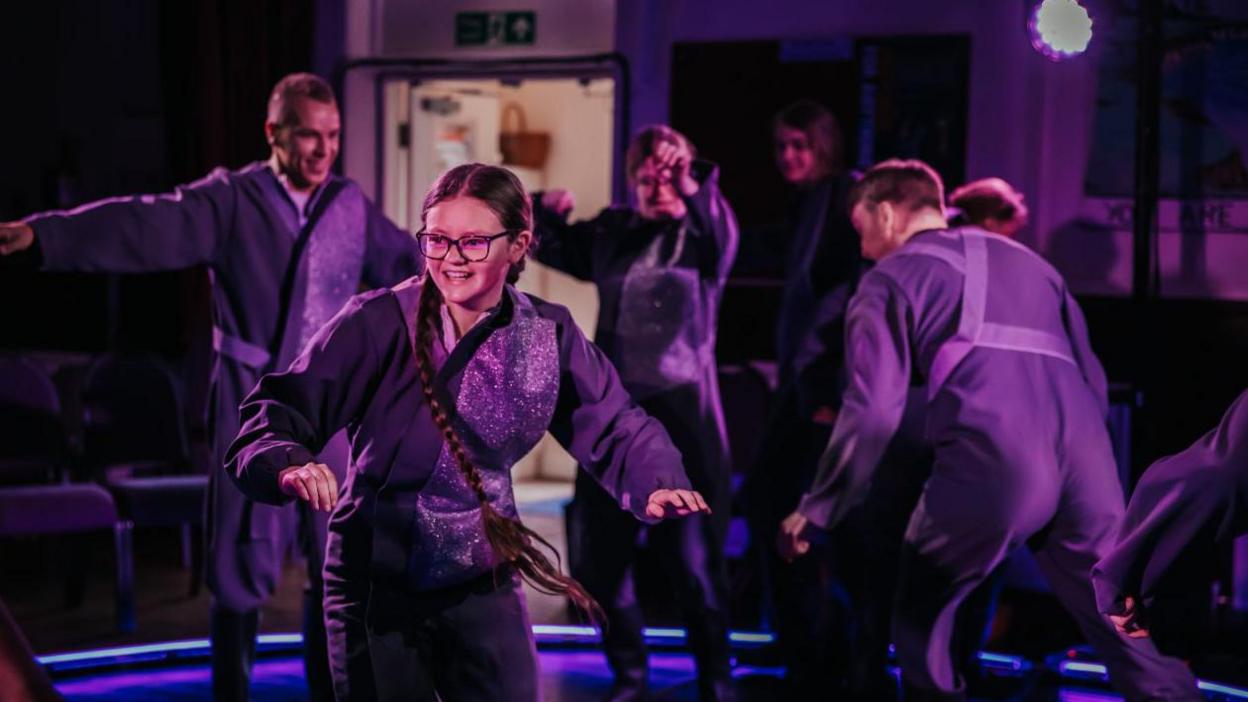 Young people make shapes on a stage lit up purple.  They are dressed in purple costumes and are all smiling.