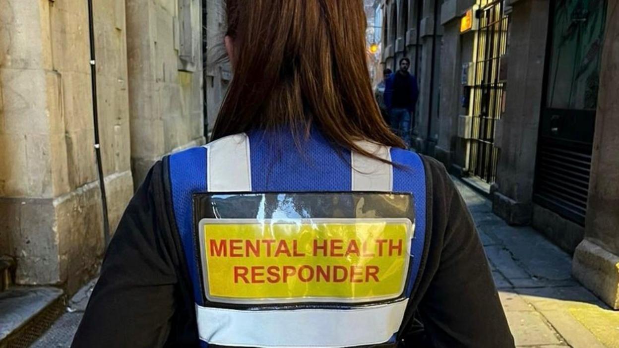 The back of a mental health responder. They are wearing a blue vest with the words 'mental health responder' written in red on a yellow background.