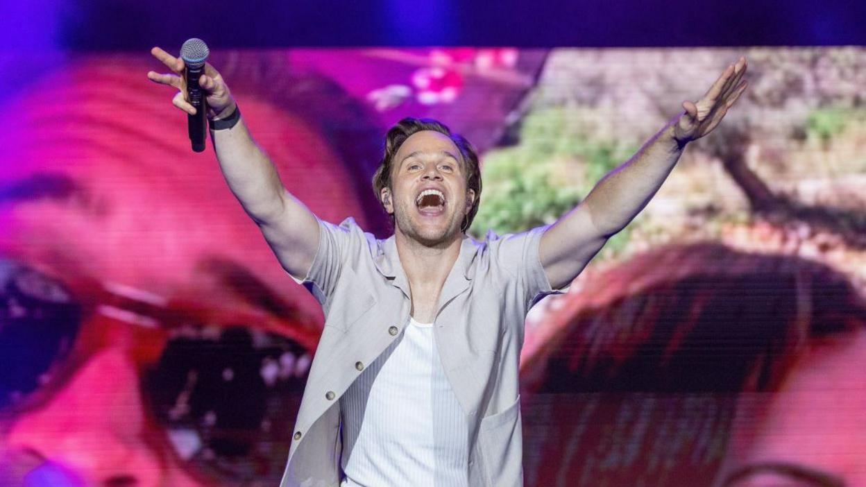 Olly Murs performing on stage in a grey open shirt with a white T-shirt underneath. He has is arms outstretched with a mic in one hand and a big smile on his face. 