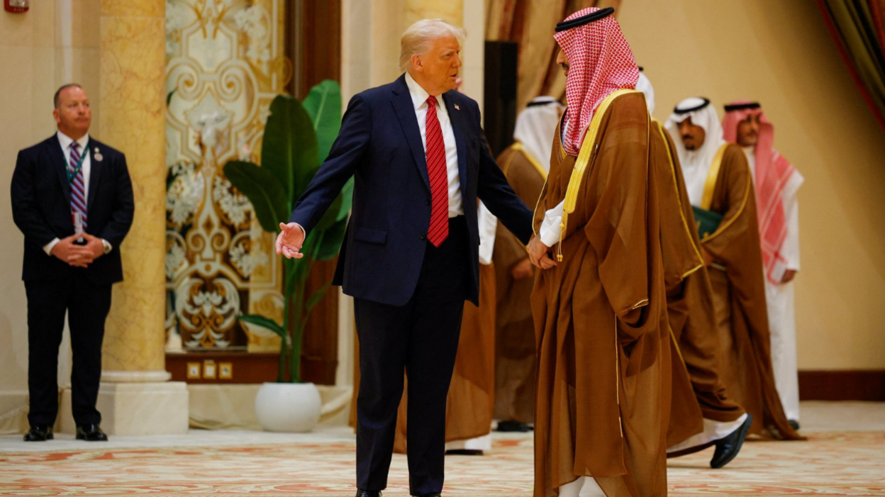 Donald Trump bën gjeste pranë Princit të Kurorës Saudite Mohammed Bin Salman. Trump vesh një kostum të errët dhe kravatë të kuqe, ndërsa princi është i veshur me një mantel të gjatë ngjyrë kafe.