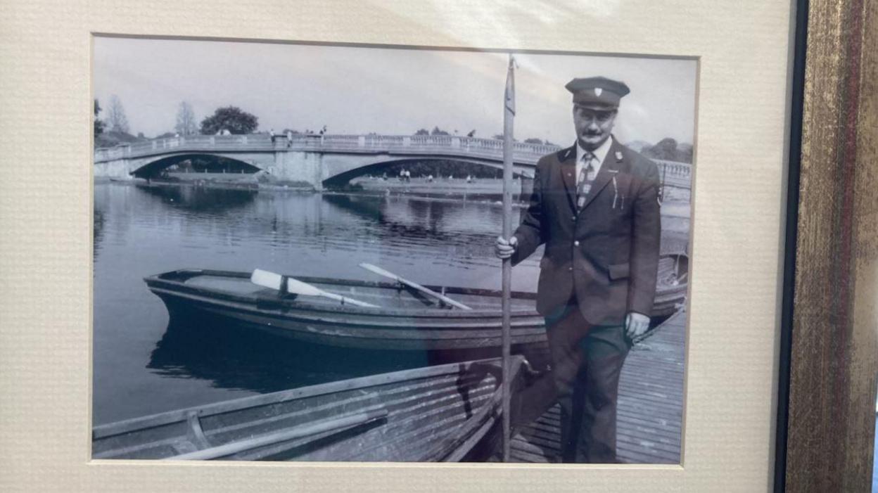 A black and white photo of Mac in a full suit and hat standing next to a lake, bridge and boats in East Park