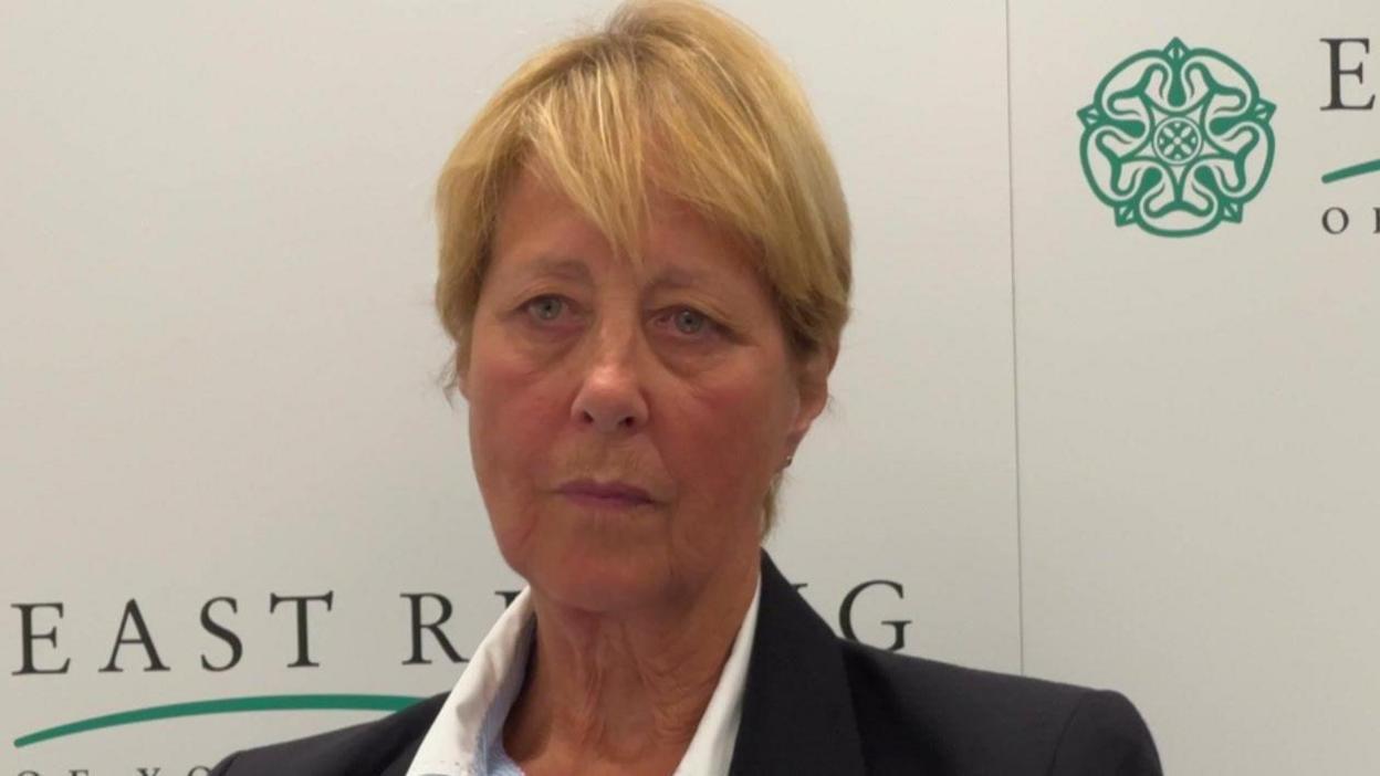 Conservative councillor Victoria Aitken - a woman with short blonde hair wearing a dark blazer and a striped shirt. She is sitting in front of a board with East Riding of Yorkshire Council's logo on it.