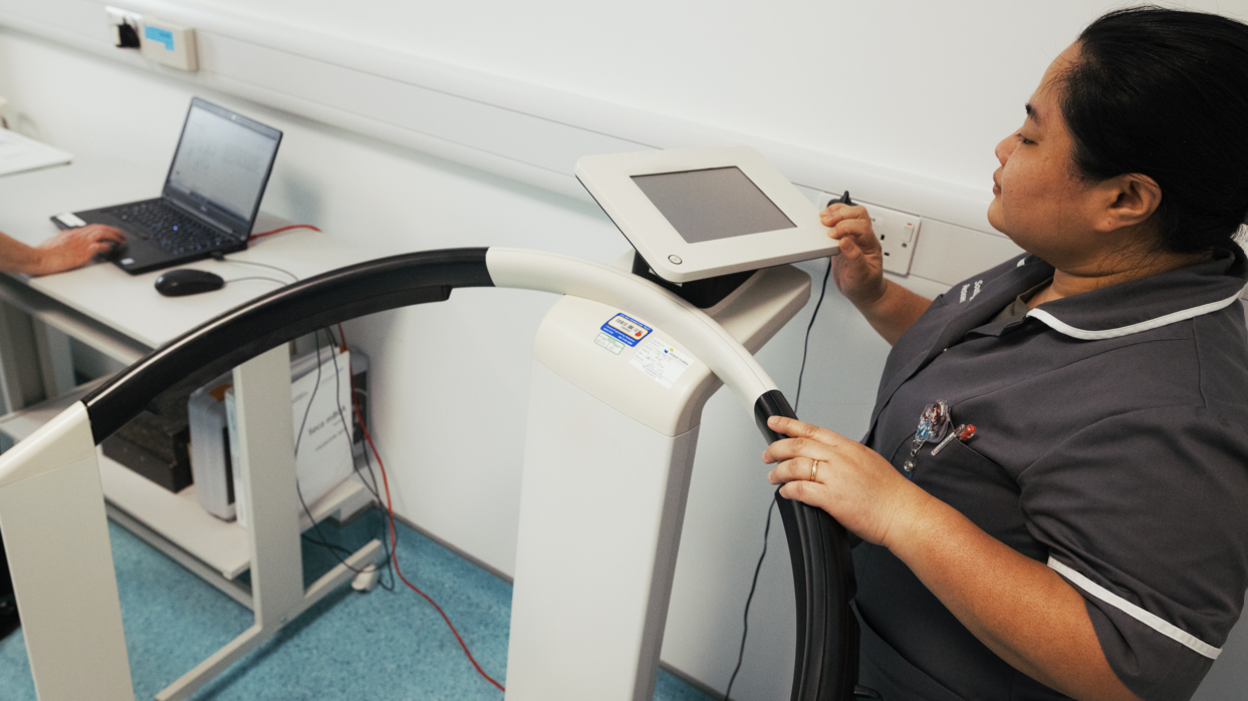 A nurse in a grey uniform working with the main body scanner at the hospital. The hand of another staff member is seen working on a laptop.