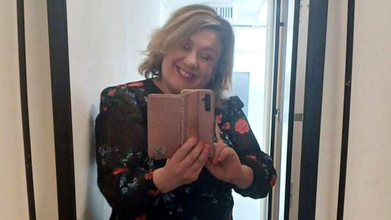 A selfie of Sarah Forrester. She is smiling at the camera and has pink lipstick on. She has shoulder-length, blonde hair and is wearing a black top with flowers.