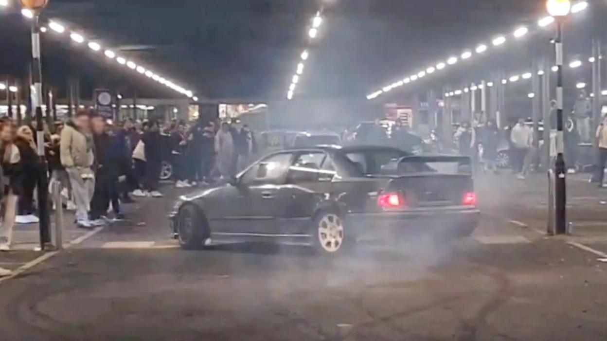 A grey car appearing to spin around doing a trick in a car park while crowds of people watch