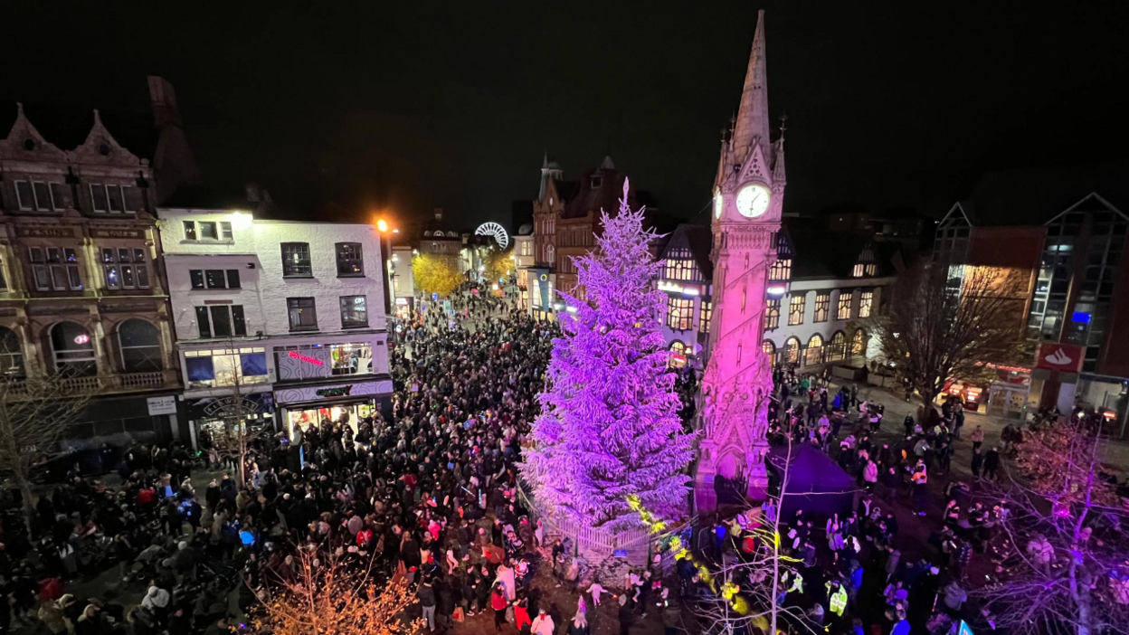 A huge Christmas tree illuminated in purple light next to a large stone clock tower