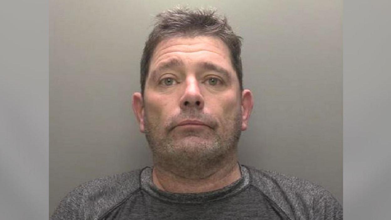 A police custody picture showing the head and shoulders of a man with short brown hair and stubble wearing a grey T-shirt against a grey backdrop.