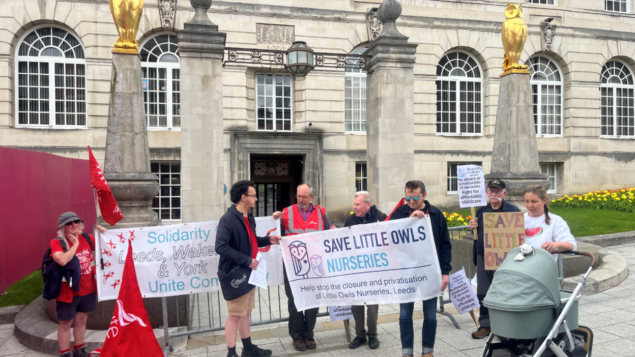Seven people hold banners reading 'Save Little Owls Nurseries' in front of a grandiose building with two gold owl statues on pillars at the entrance. A mother with a pram can be seen on the right-hand side.