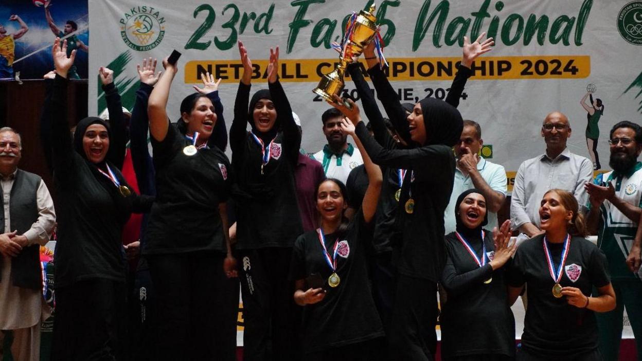 The Cosmopolitan Roses raising the trophy as they cheer and clap. Smiling officials look on.