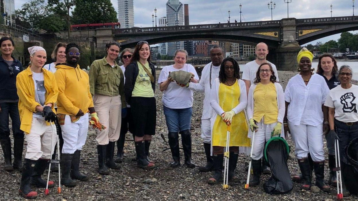 A group of people stand on the foreshore of the River Thames in front of Vauxhall Bridge/ They are wearing wellies and some have litter pickers in their hands.