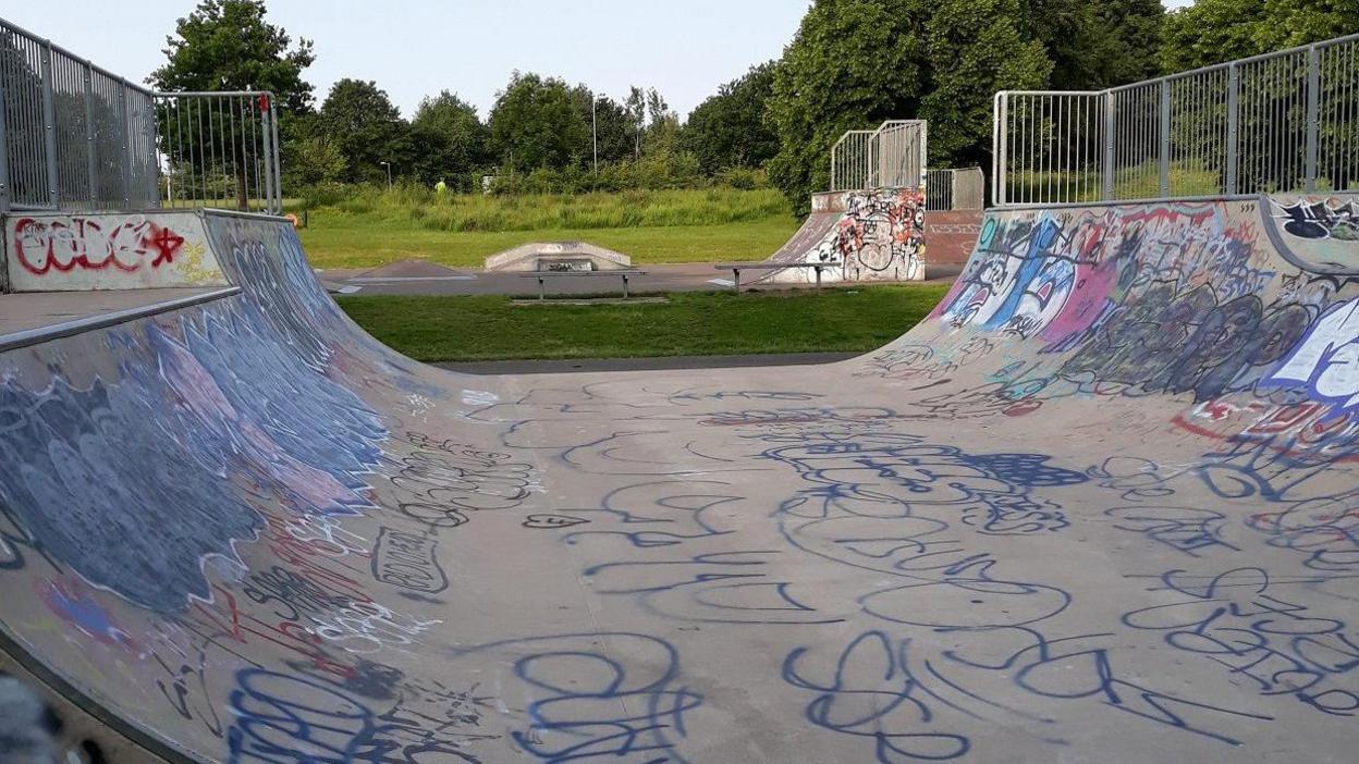 The concrete half-pipe from the side in a skate park with the sides curving upwards either side. There is grass behind and a further half-pipe in the distance. Graffiti is scrawled all over the concrete.