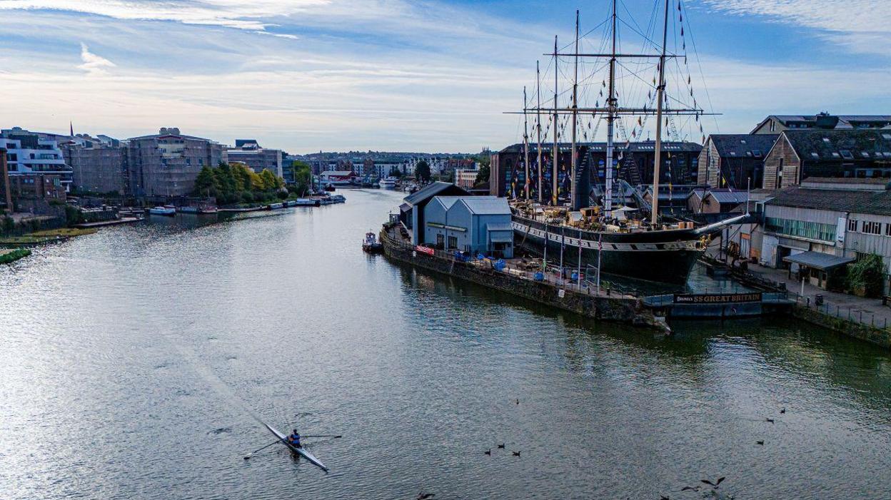 A rower passes the SS Great Britain on Bristol's harbourside. There are also gulls visible on the surface of the water. It is taken just after sunrise.