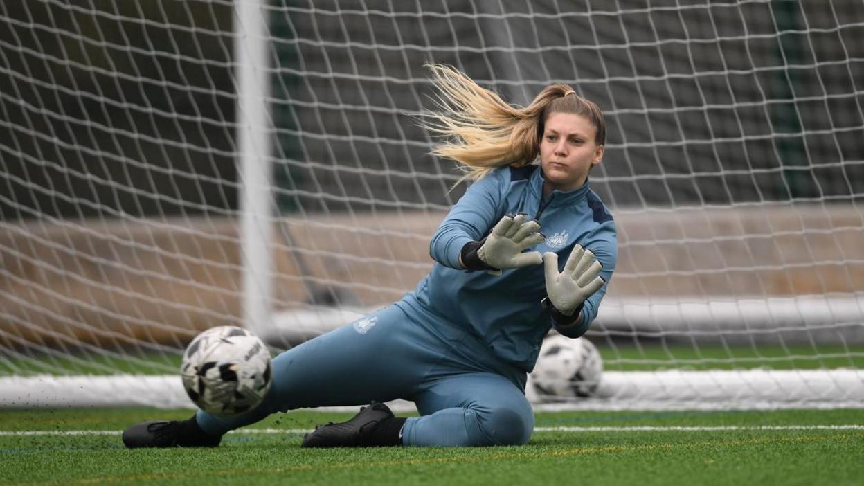 Grace Donnelly saving a goal during a training session. She has dived towards an on-coming ball and has both palms up ready to deflect it. She is wearing her blue training tracksuit.