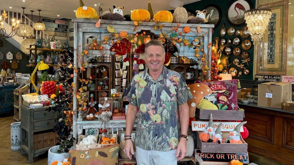 A man with grey hair in a multi-coloured shirt is smiling at the camera in front of many household and gift items in a store.