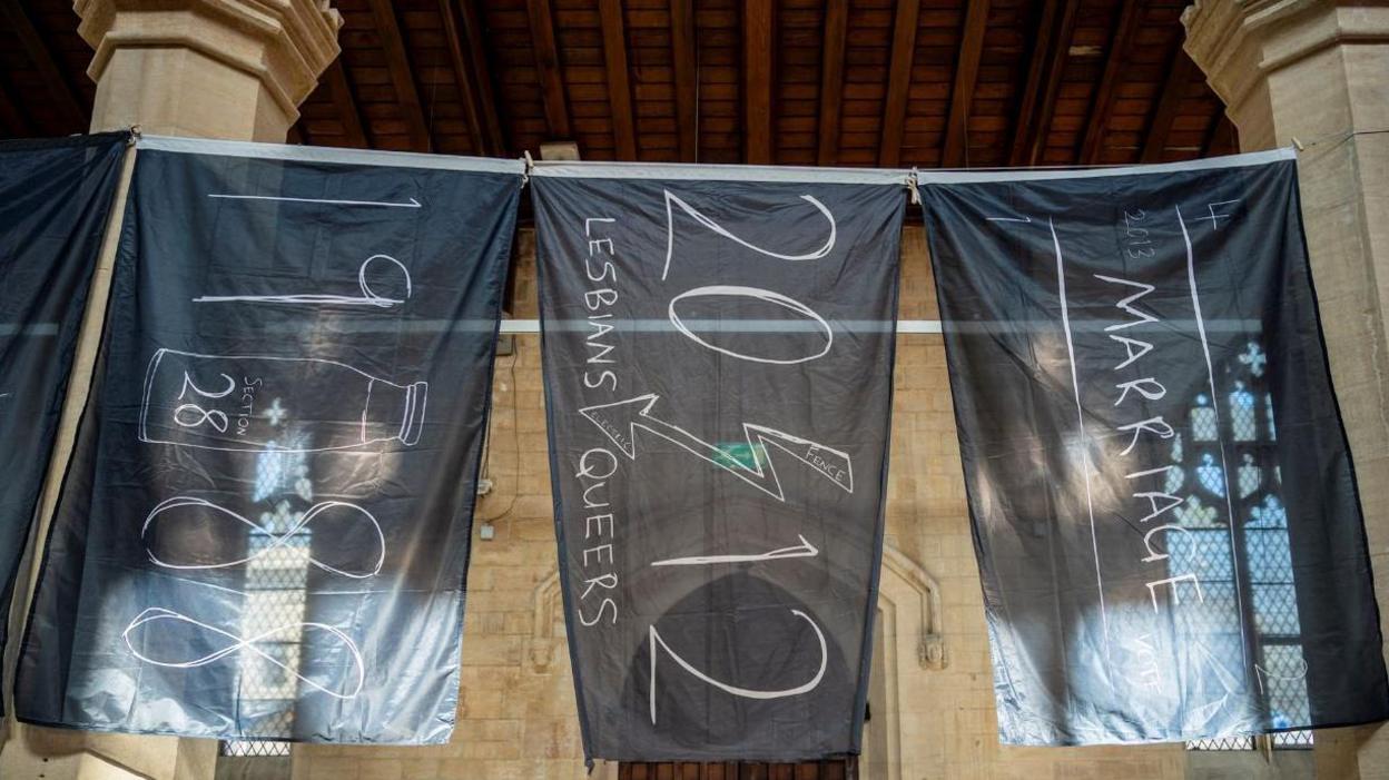 Three black flags draped down a wall, with various messages written on them in white, including one that says "1988" with "Section 28" in a bottle.
