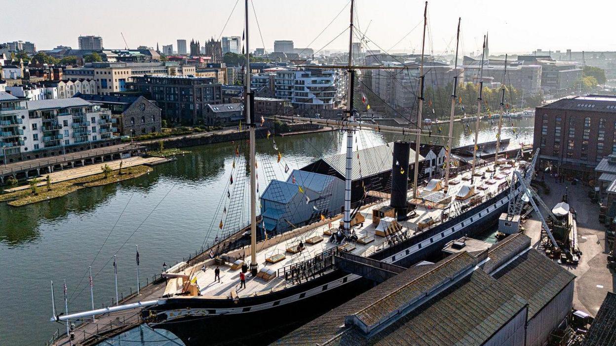 An aerial image of the SS Great Britain in its dry dock in Bristol. The ship is in the foreground and the city centre is visible in the background. The sun is shining on the ship's deck and the waters of the harbour are still