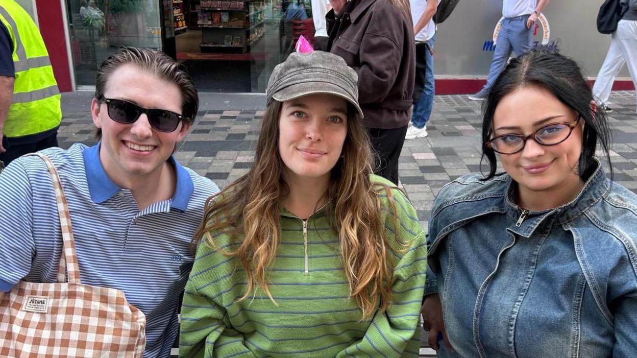 A man and two woman on a bench in Glasgow. The man is wearing sunglasses and a striped top. The woman in the middle is wearing a green striped fleece and hat and the woman on the right is wearing a denim jacket and glasses. 