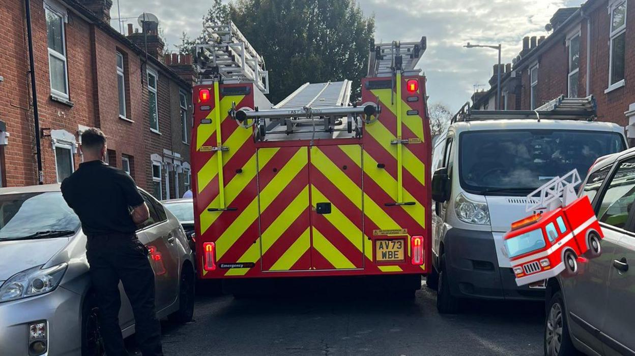 A rear view image of a fire engine. It is squeezing past parked cars as a male firefighter, dressed in black looks on. 