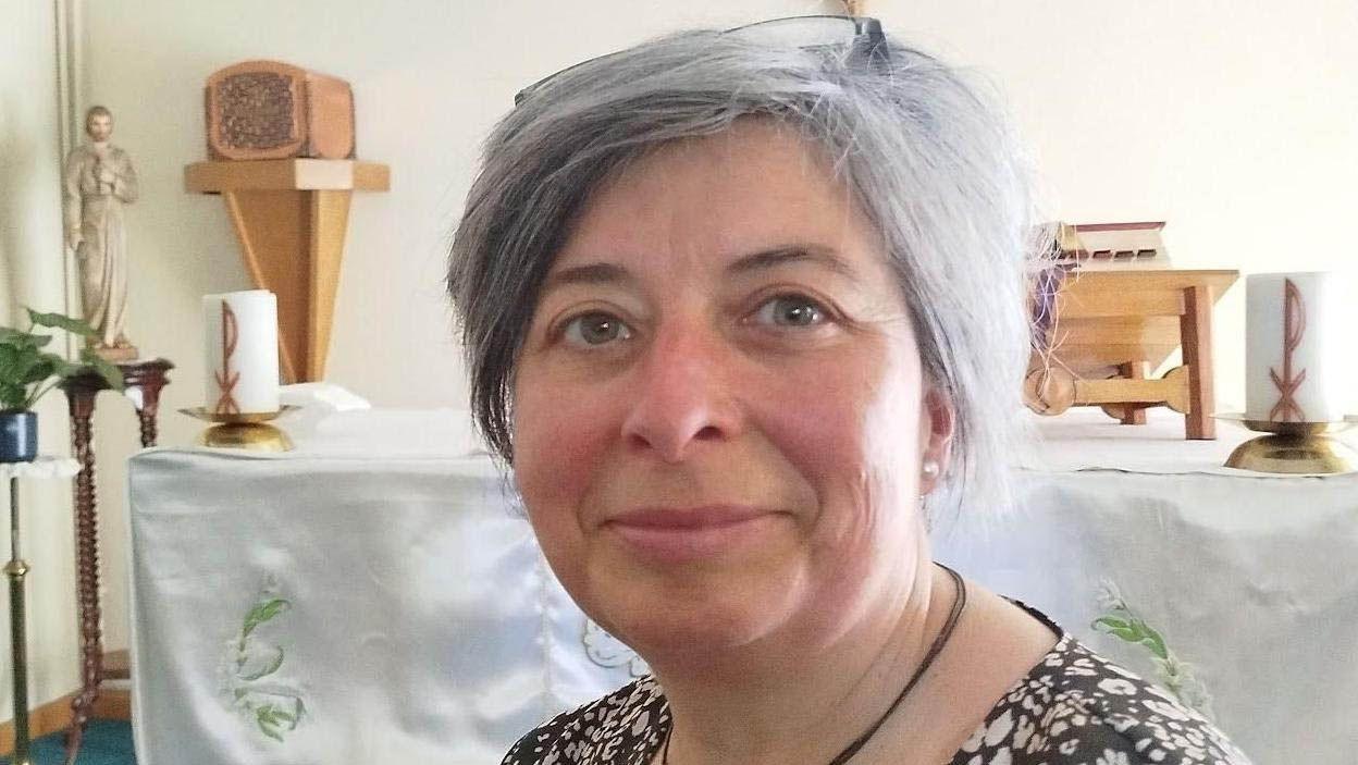 Head-and-shoulders shot of Eva Ripoll Breau, sitting in front of an altar and smiling. She has short bobbed grey hair and wears glasses on top of her head.