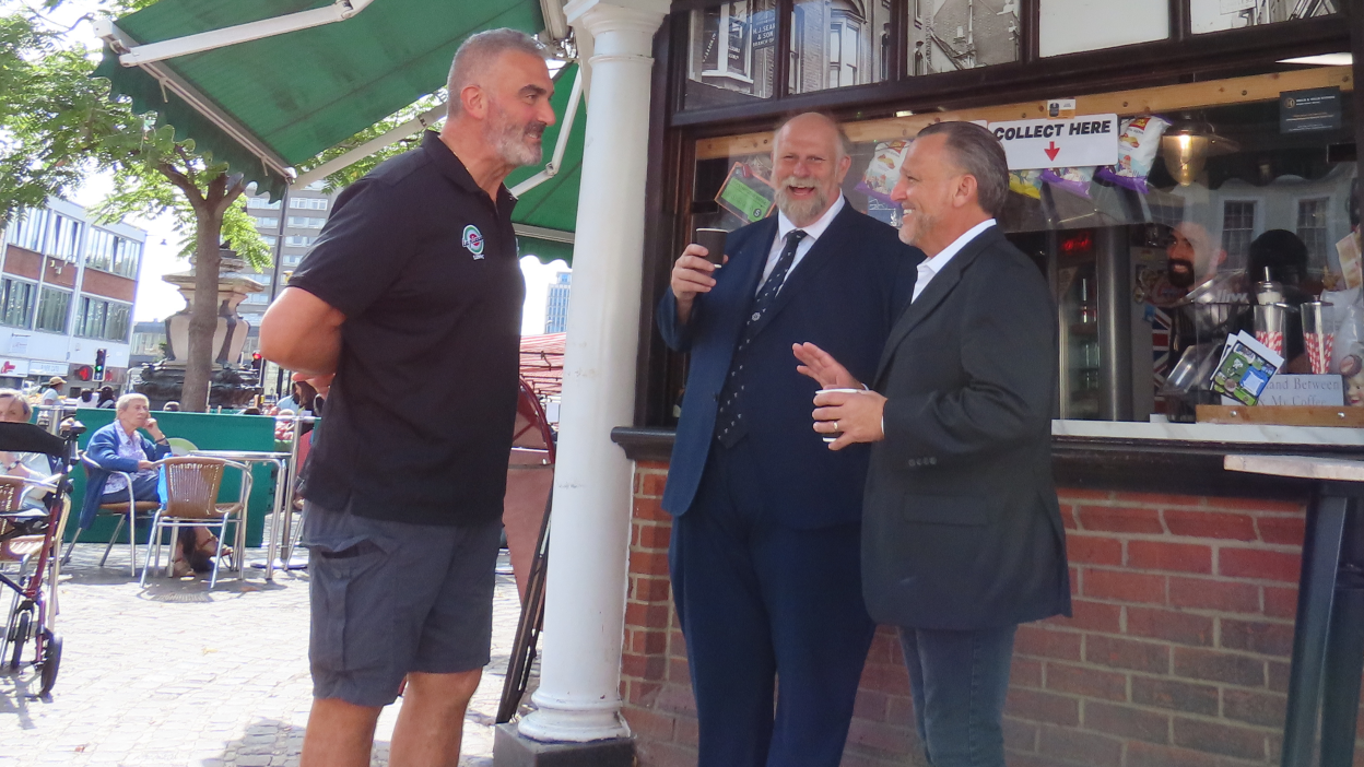 Libby Lionetti, standing in front of his cafe, talking to Mayor Tom Wootton, holding a coffee cup and John McReynolds