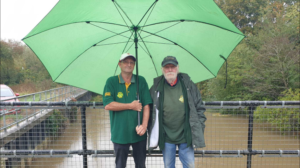 Nigel Russell (left) and chairman Peter Gilbert, standing on a bridge over the river. Nigel is holding a very large green umbrella, which they are both sheltered under and the river, which is a light brown colour, can be seen running behind them. 