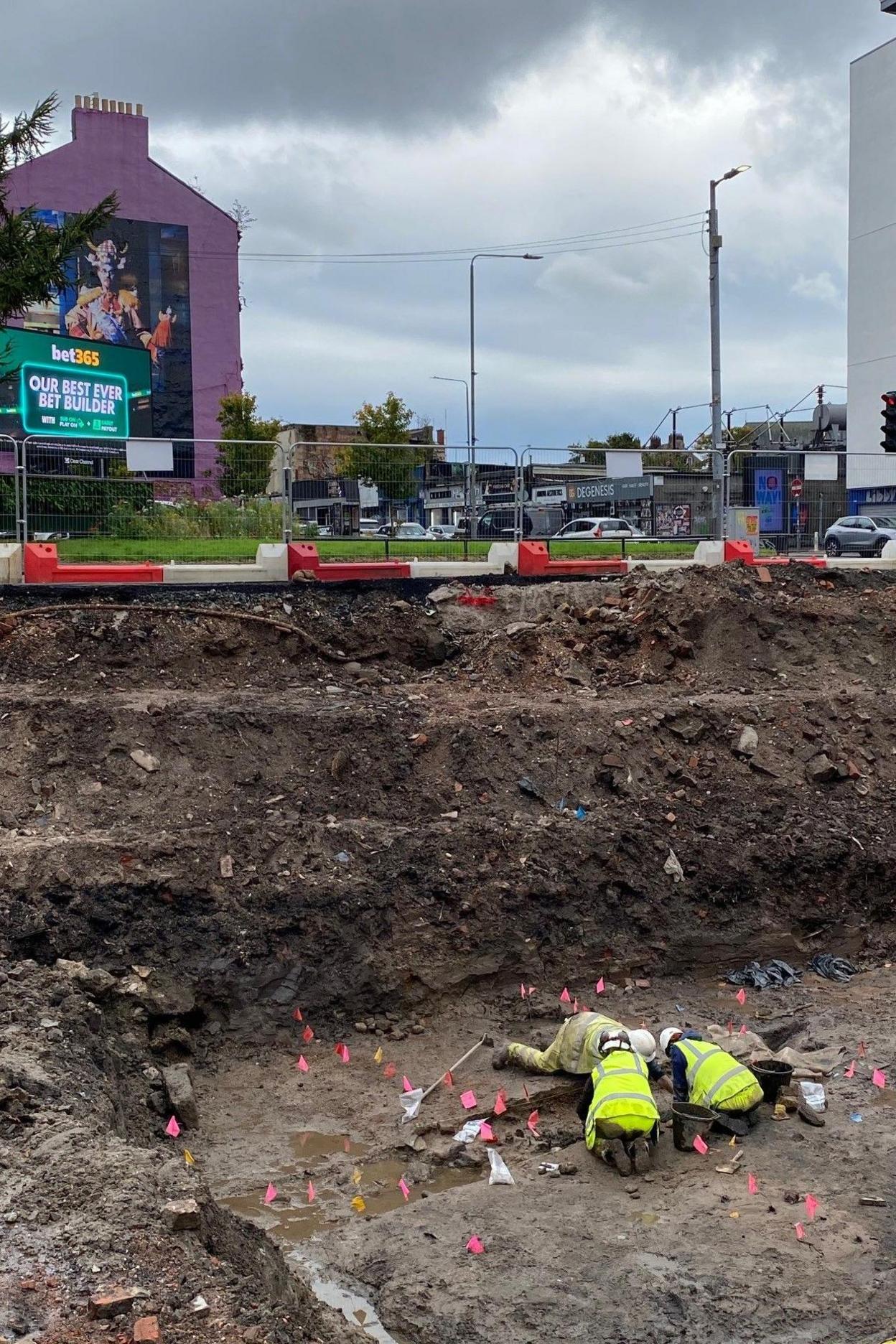 An archaeological dig with archaeologists on the ground digging in some trenches. They are wearing high vis jackets. Above ground level is a building with a mural of Billy Connolly on its side