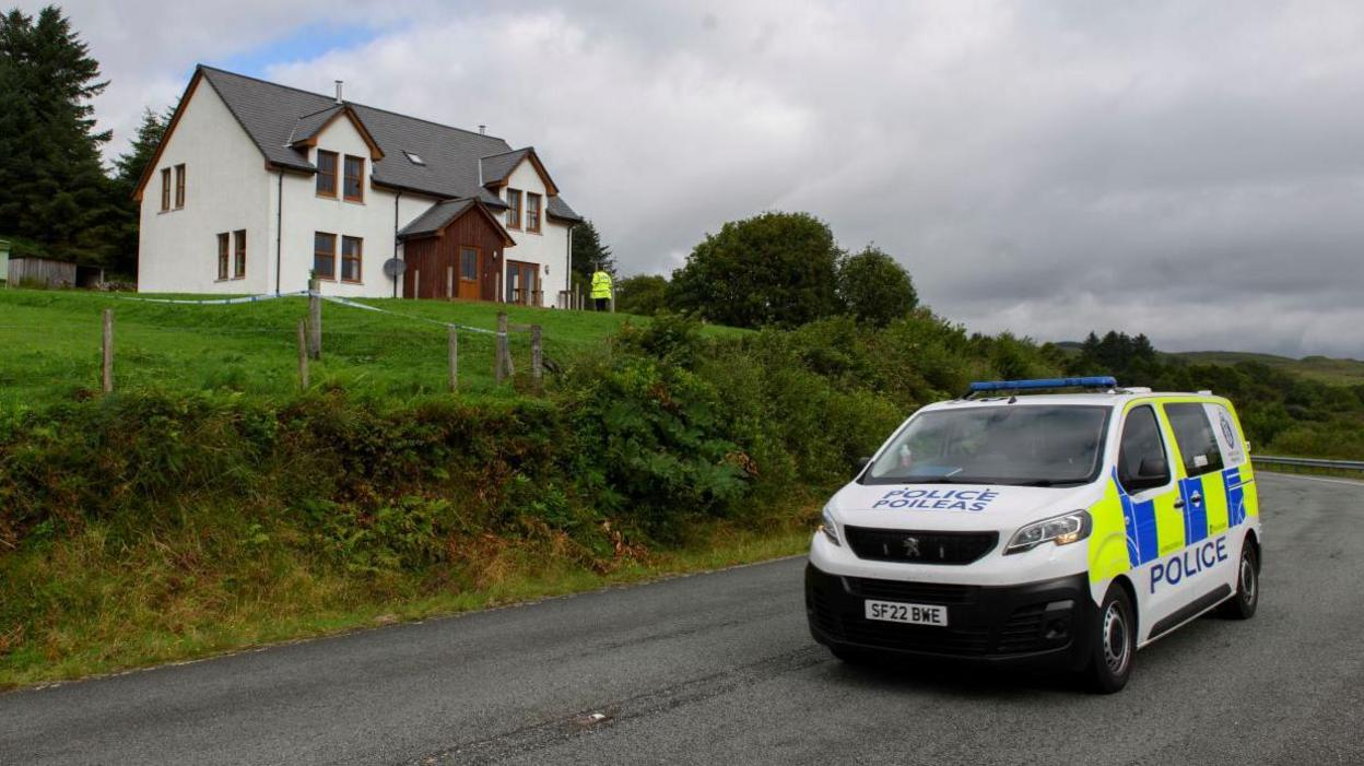 A police van is parked on a road below the two-storey house. There is a police office standing near front door of the property.