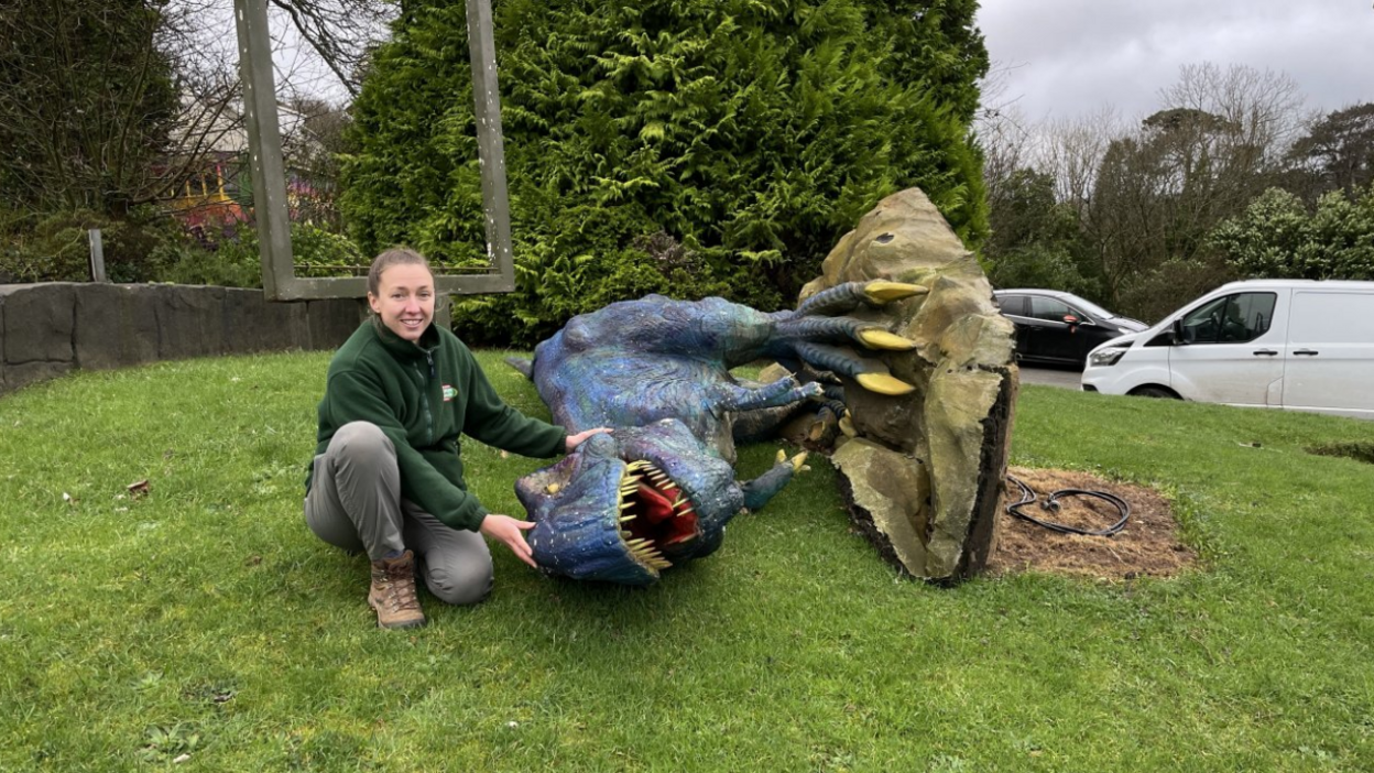 Louisa Bartlett crouched beside a damaged T-Rex figure. She is wearing a green fleece jumper with grey trousers. The T-Rex is blue with yellow claws. They are on grass.