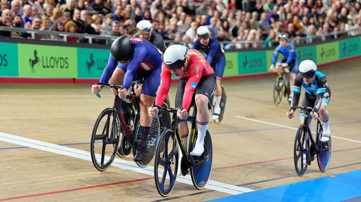 Ioan leading a tight pack of cyclists at the velodrome. They are all wearing black and white helmets and cycling Lycra, Ioan wears a red top and black shorts. A crowd can be seen in the background.