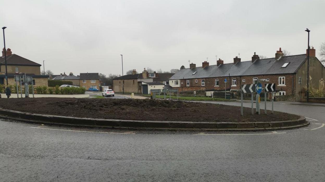 A roundabout that is covered in bare soil. Houses line the road leading up to the roundabout.