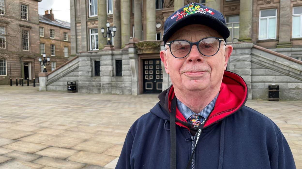 Jerry Williams is stood in front of Birkenhead Town Hall. He has grey hair and is wearing dark rimmed glasses and a blue cap. He is wearing a blue shirt and multi-coloured tie and a blue zipped hooded top.