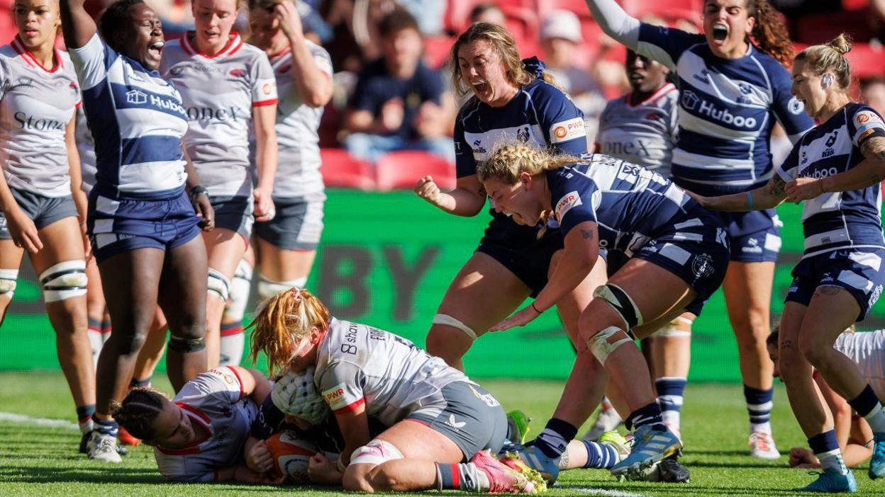 Bristol Bears Women players celebrate with shouts and clenched fists as Lana Skeldon scores their second try against Saracens at Ashton Gate. Skeldon is being held by two Saracens players but she has managed to ground the ball