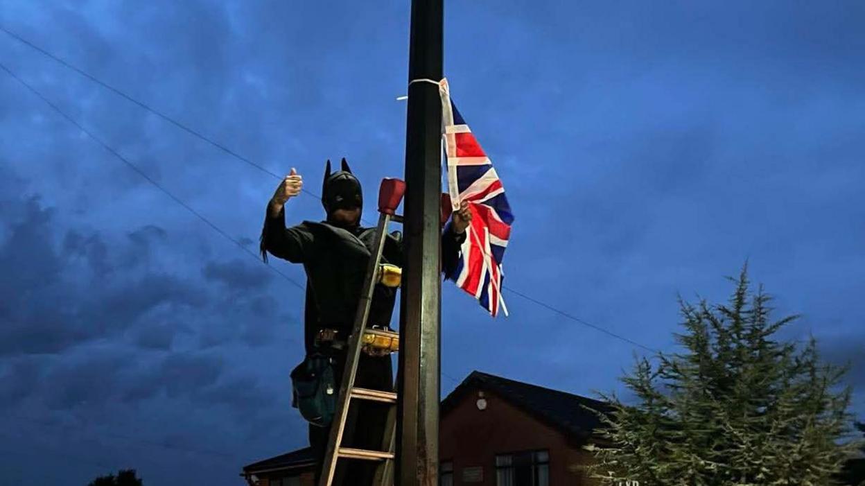 Man in black Batman suit gives a thumbs-up as he ties a Union flag to a street post on a cloudy evening.