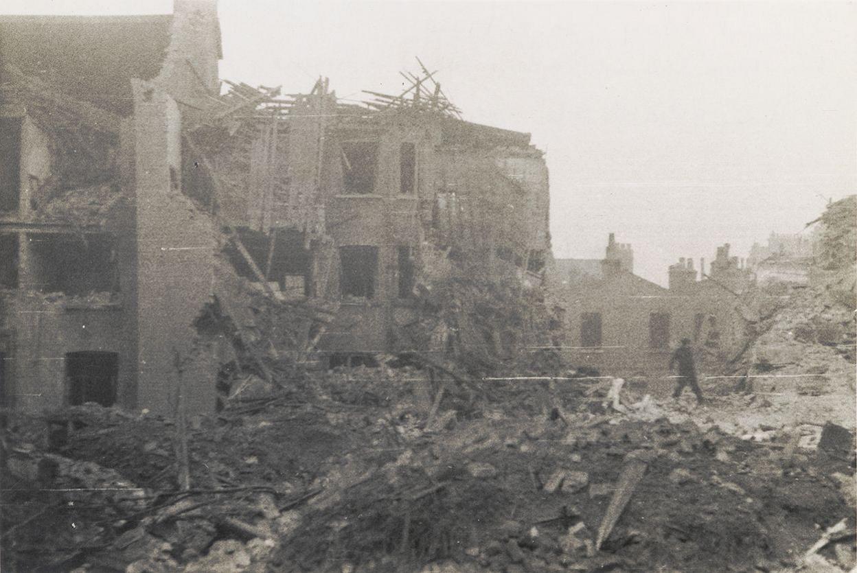 Black and white photo of a man walking beside bomb damaged homes with rubble piled up beside several damaged houses