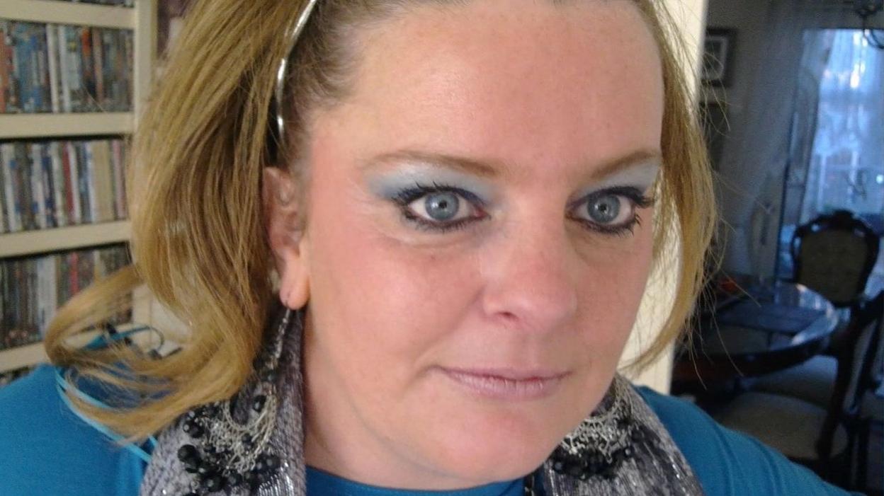 Jodey Whiting is looking towards the camera. She has blonde shoulder-length hair with a silver hairband and is wearing a a silver coloured scarf and a light blue jumper