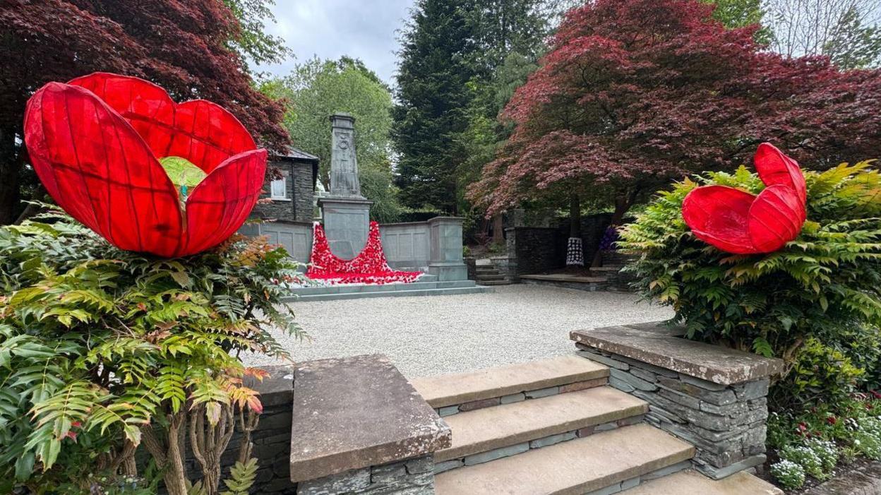 Two giant knitted poppies appear either side of bushes and a stone entrance, leading to a war memorial which has smaller, knitted poppies cascading down from it.