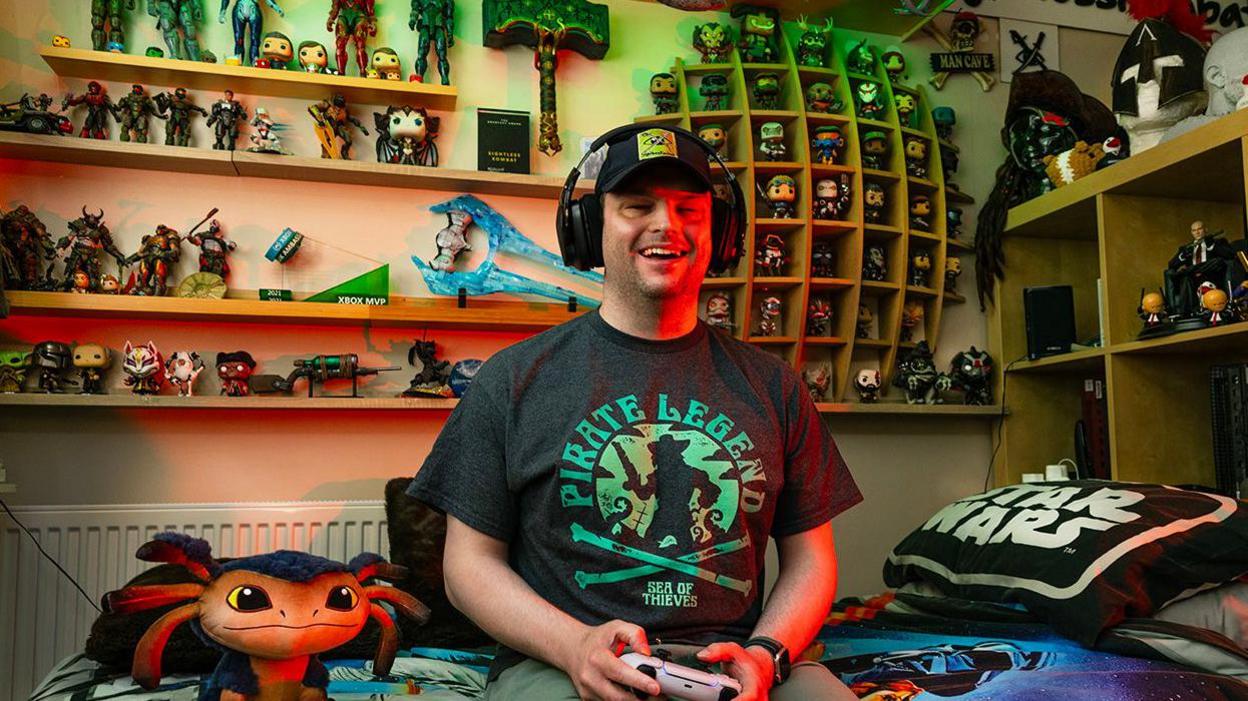 A man wearing headphones, sat on a bed holding a games console remote. He has a black "pirate legend" t shirt on, and green shorts. Behind him are shelves filled with character statues.
