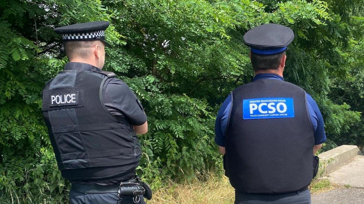 Two officers in uniforms and black hats stood facing forwards. On the backs of their uniforms one reads 'POLICE' the other reads 'PCSO' stood in front of police taper by a bridge in a leafy area.