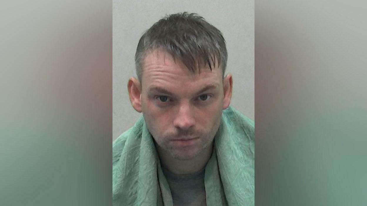 Mugshot of a man with brown hair and wrapped in a green blanket