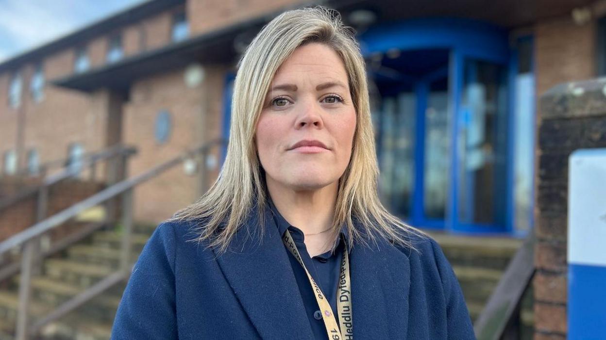 Katie Cuthbertson mewn cot las a lanyard Heddlu Dyfed Powys yn sefyll o flaen pencadlys yr Heddlu