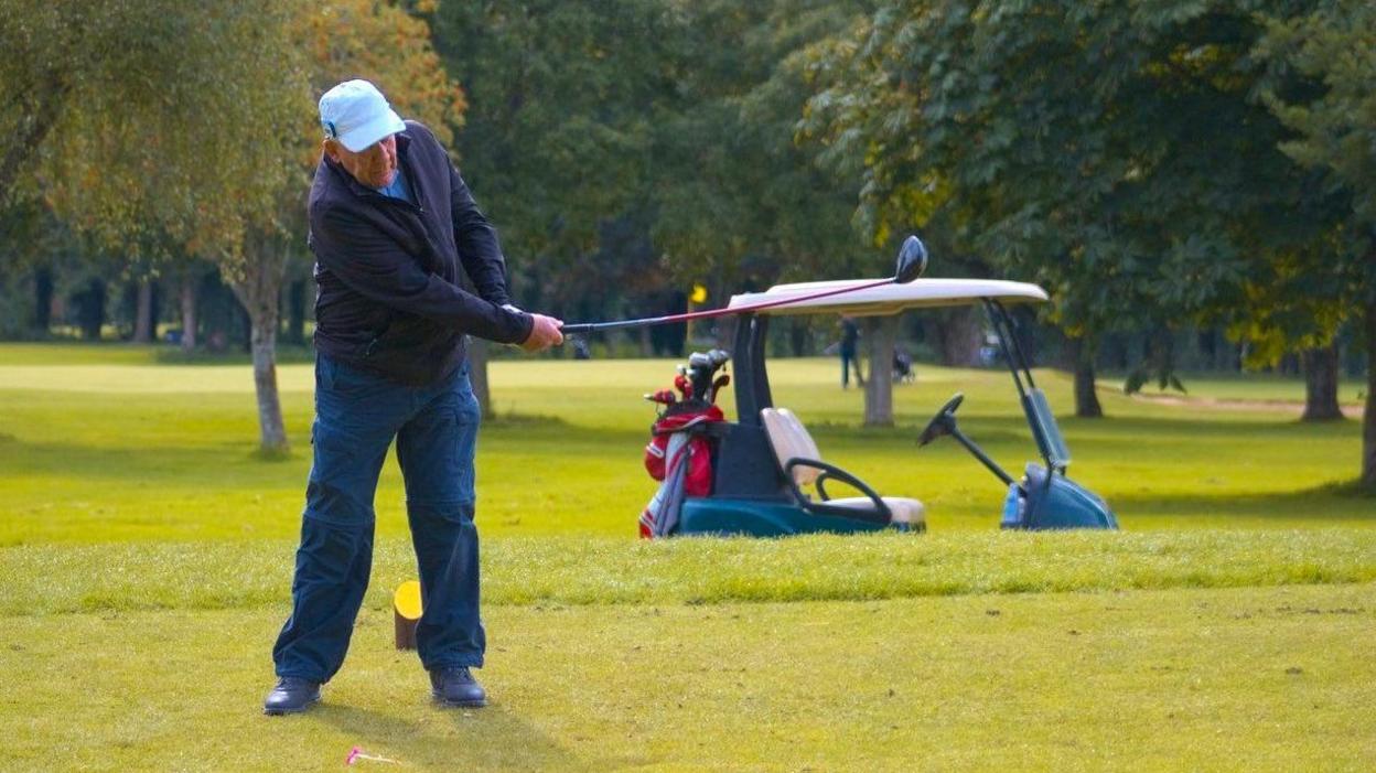 Danny Daniels, who is wearing blue golfing gear and a light-blue baseball cap, swinging his golf club on British Blind Masters golf course at Meon Valley. There are trees, greens and a golf buggy behind him