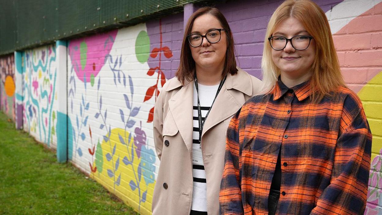 Two women are stood in front of a wall which has colourful floral patterns painted on it.