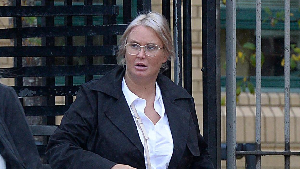 A woman, with blonde hair, wearing a white blouse, black jacket and glasses. She is looking to the left of the photo. There are black and grey gates behind her. 