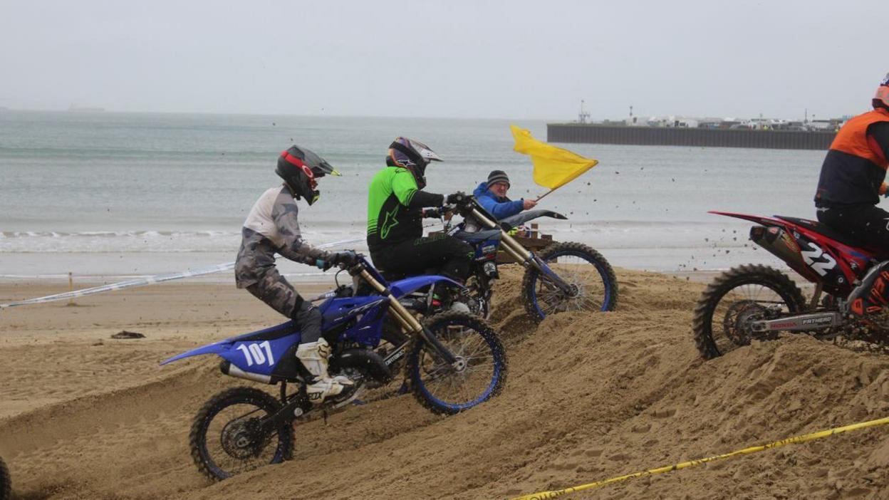 Man waving a yellow flag as riders overcome a sand obstacle. The sea is behind them. It's cloudy.