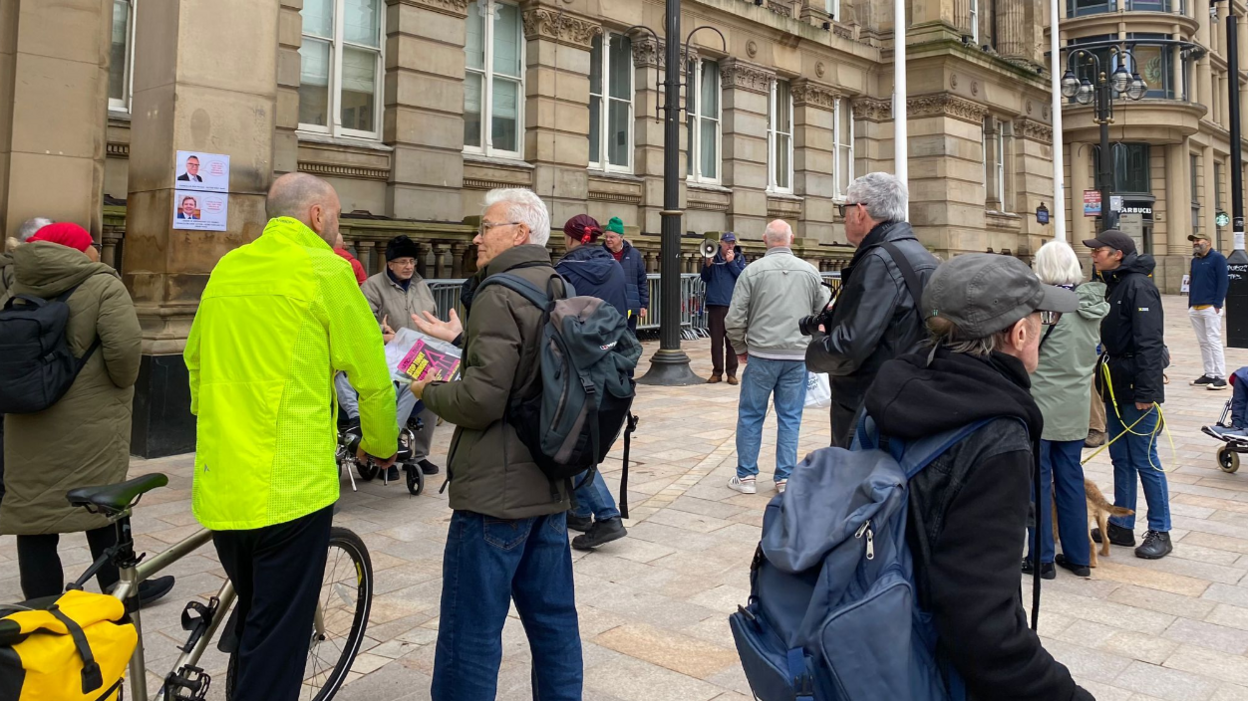 Men and women stand outside the council house entrance, some with leaflets in their hands. One man in a fluorescent jacket holding a bike's handles is standing talking to another man. There is another man with a dog.  