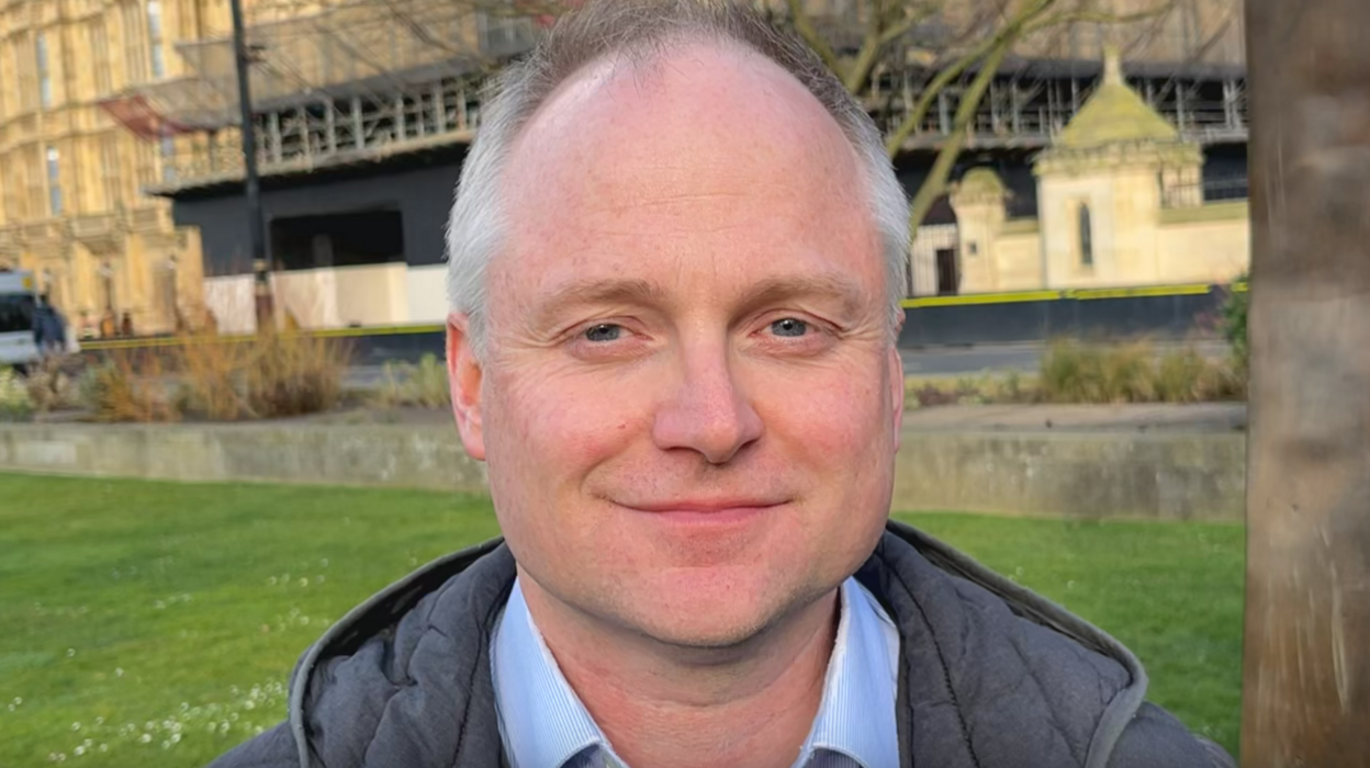 Andy Statham is smiling at the camera outside the Houses of Parliament. He is wearing a grey coat and light blue shirt. 