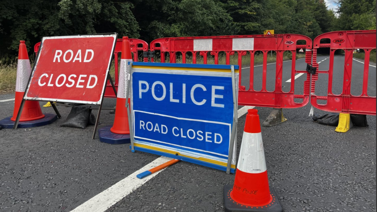 A blue and white police road closure sign stands next to a red and white one with traffic cones in between and red plastic barriers behind on a tree-lined road