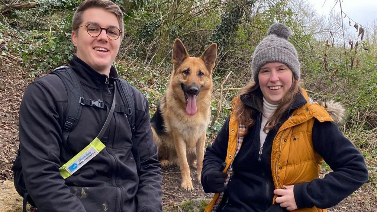 Adam Drysdale and his partner, Elle Manners. Both of them are pictured outside, sitting on a rock with a German Shepherd guide dog sat in between them with its tongue out. Adam has short brown hair and glasses. He is wearing a black coat and smiling at the camera. Elle is wearing a grey bobble hat and a mustard coloured gilet. 